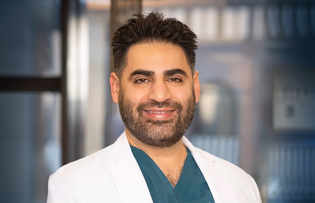 Smiling male doctor with dark hair and beard wearing a white coat over green scrubs in a blurred medical office background.