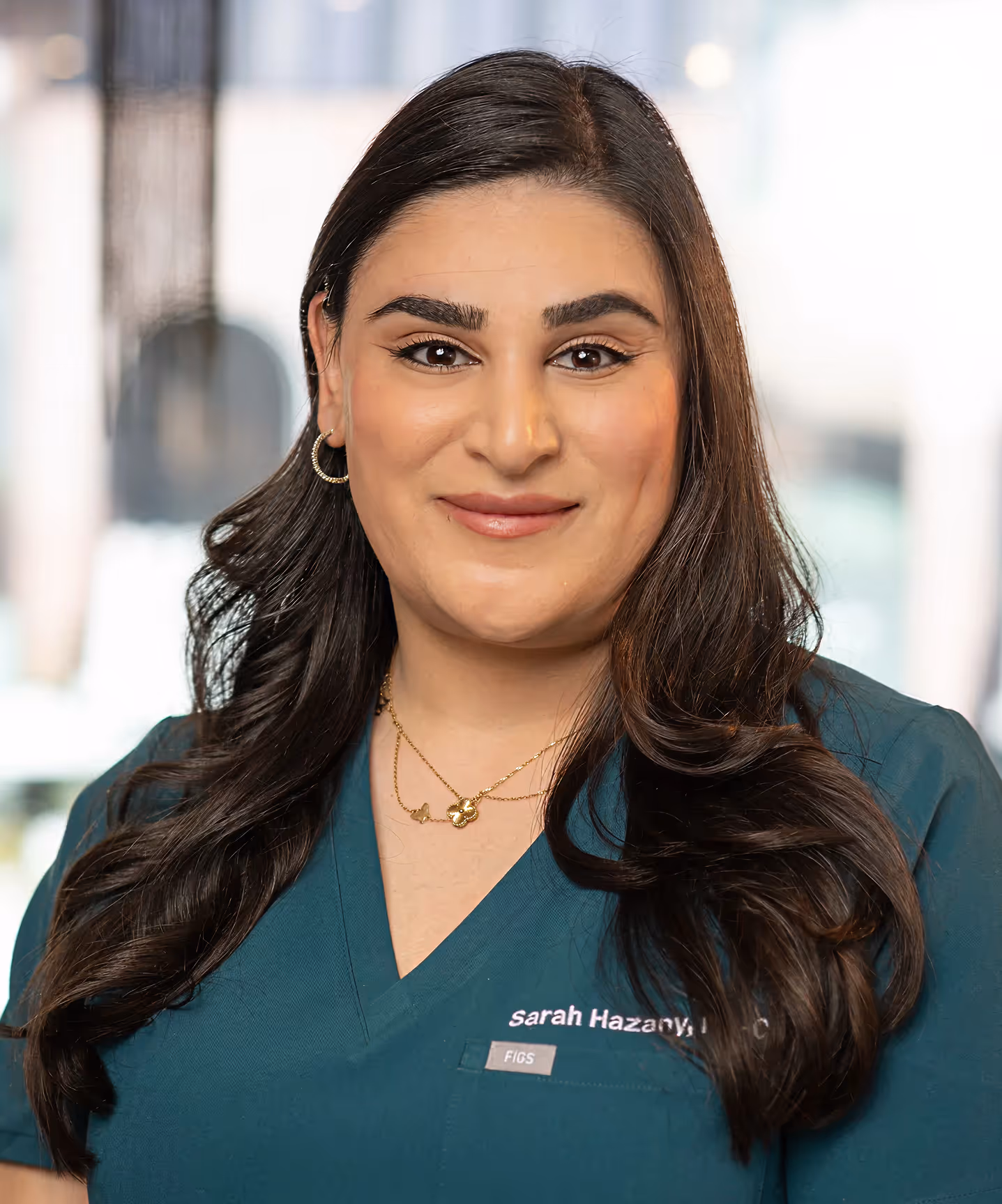 Smiling woman in teal medical scrubs with long dark hair and gold necklace in a softly blurred indoor setting.