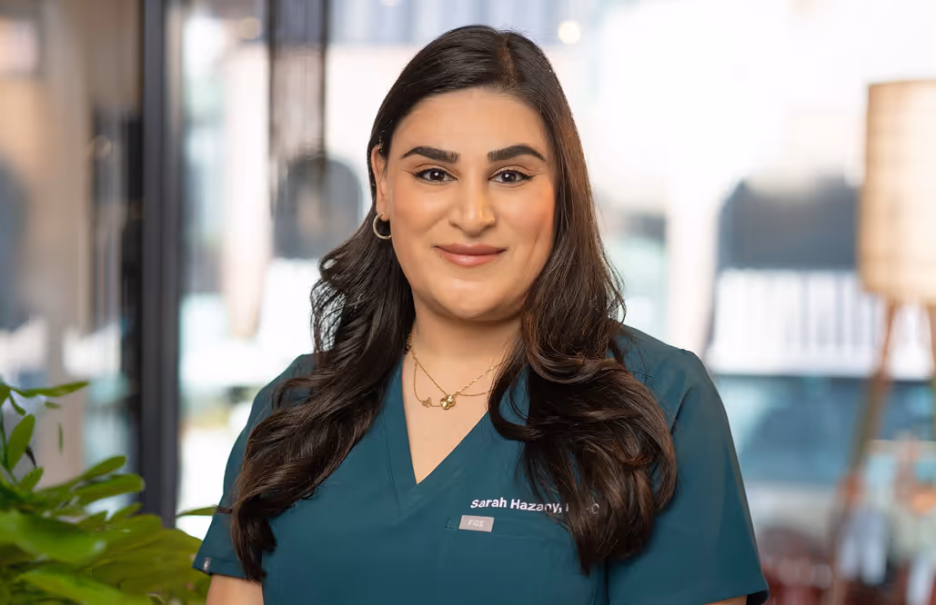 Smiling woman with long dark hair wearing green medical scrubs and standing indoors.