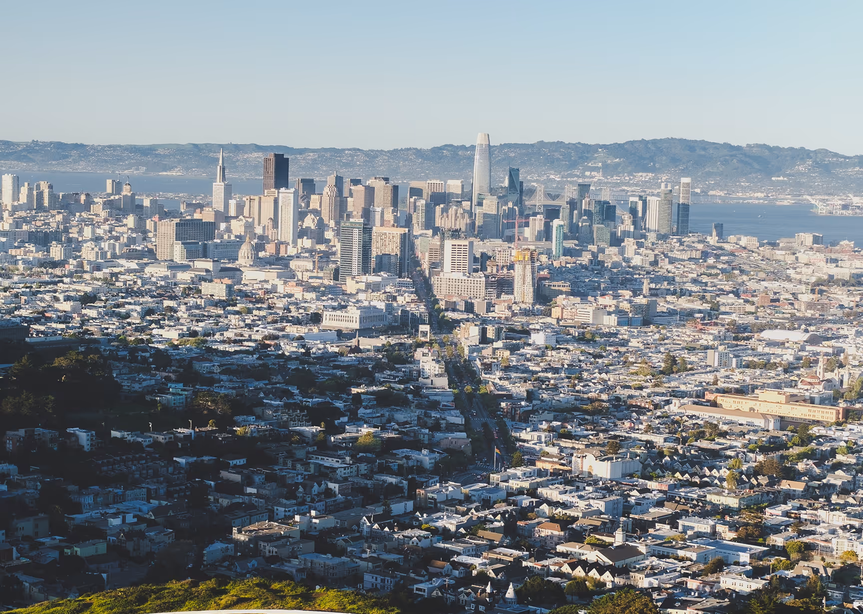 Aerial view of downtown San Francisco with dense urban buildings, prominent skyscrapers, and the Bay Bridge visible in the background.