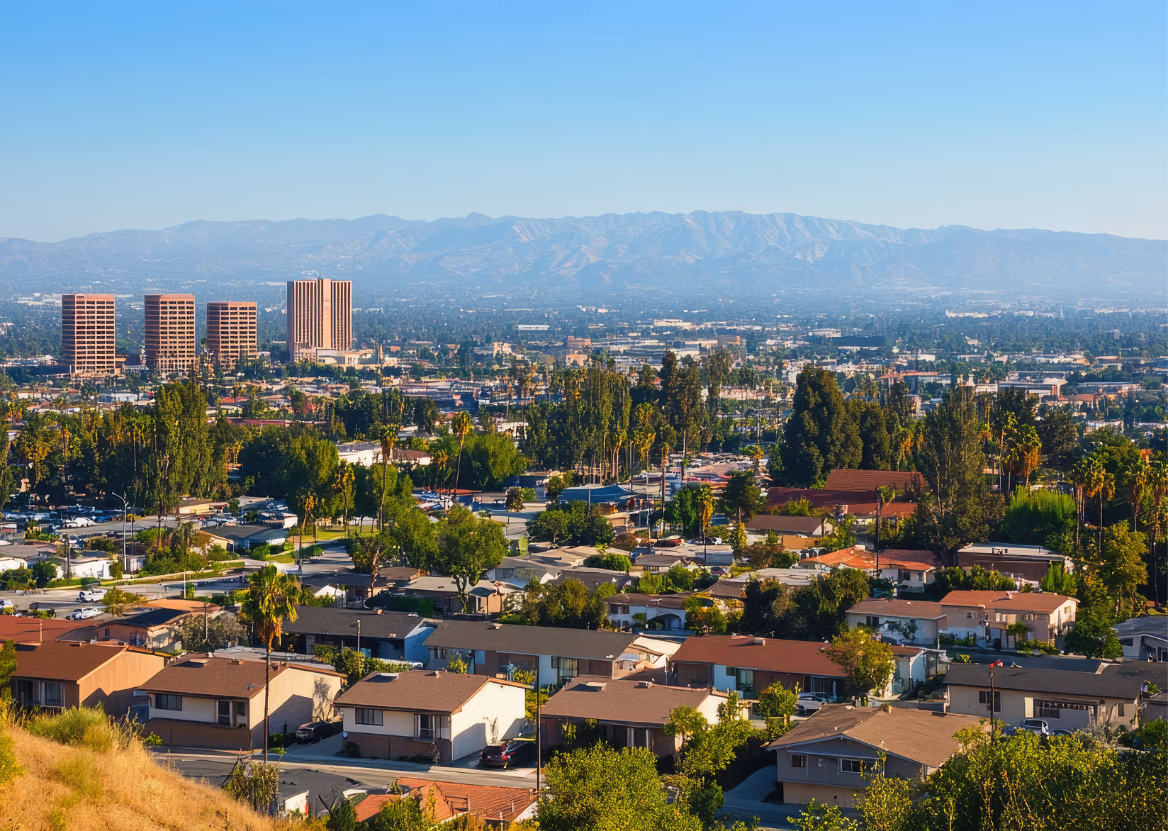 Suburban neighborhood with single-story houses and trees in the foreground, tall office buildings in the midground, and mountains under a clear blue sky in the background.