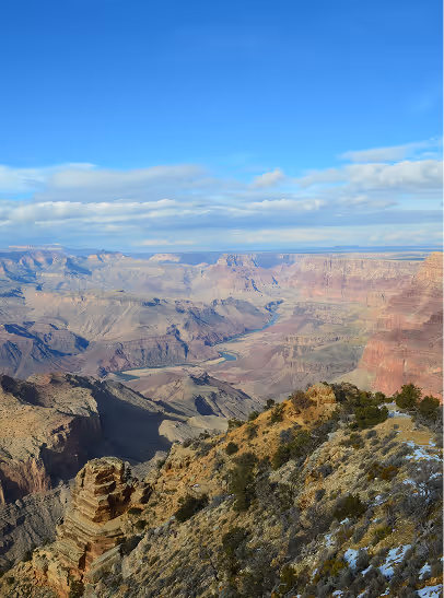 View of the Grand Canyon with layered rock formations and the Colorado River winding through the valley under a blue sky with scattered clouds.