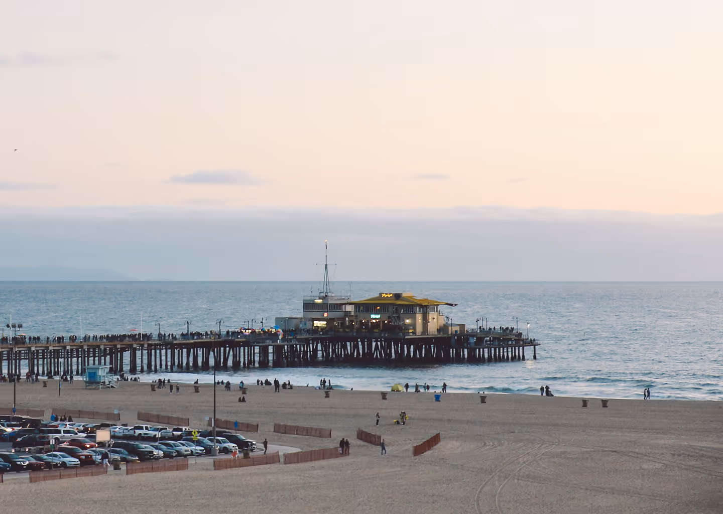People walking on a pier extending into the ocean at sunset with a building at the pier's end and a sandy beach with parked cars in the foreground.