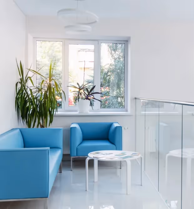 Modern waiting area with blue armchairs, white round table, large window, and green potted plants.