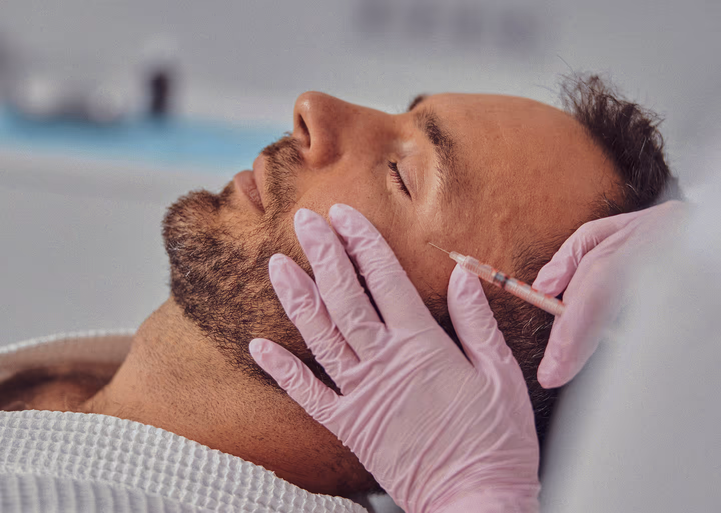 Man with closed eyes receiving a cosmetic injection in his cheek by a gloved hand.