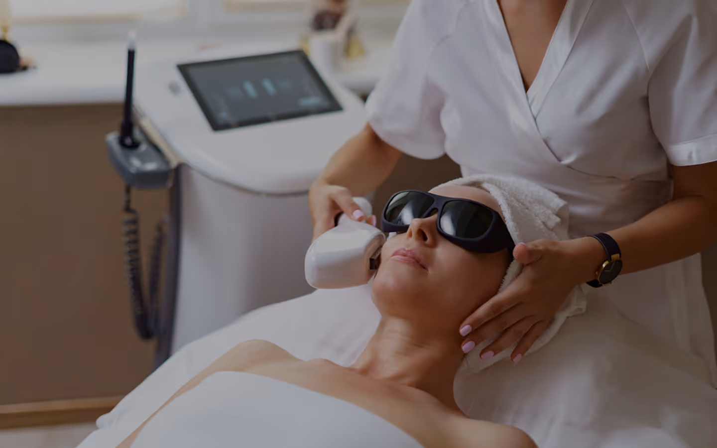 Woman wearing protective goggles receiving a facial laser treatment from an esthetician in a spa setting.
