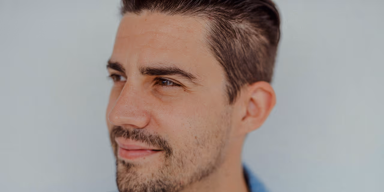 Close-up of a man with short dark hair and light facial stubble looking to the left against a plain light background.