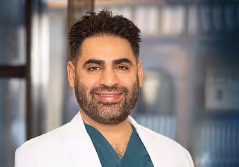 Smiling man with dark hair and beard wearing a white coat and green scrubs in a professional setting.