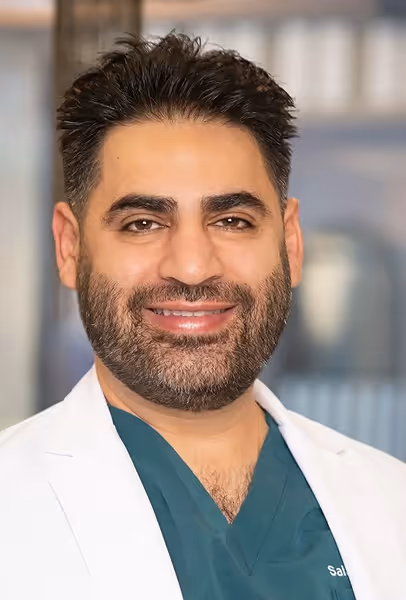 Smiling man with dark hair and beard wearing green medical scrubs and a white coat.