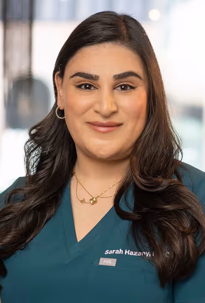 Portrait of a woman with long dark hair wearing teal medical scrubs and gold hoop earrings, smiling.