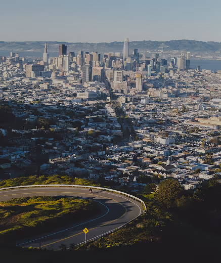 Curving road on a green hillside with a cyclist and the San Francisco city skyline in the background under a clear sky.