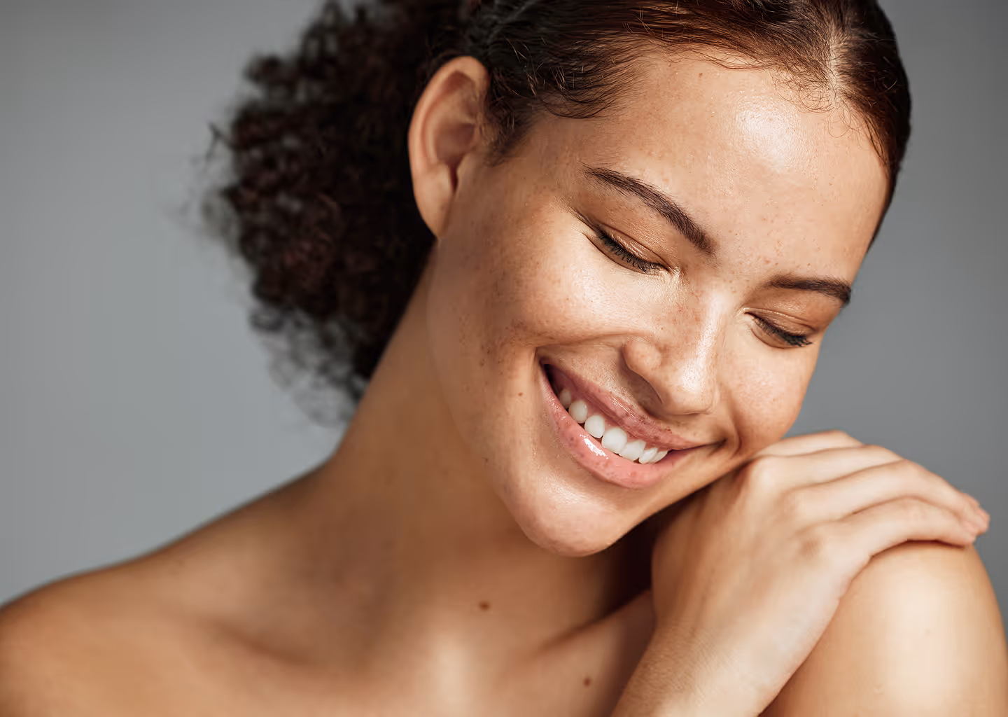 Close-up of a smiling woman with natural curly hair resting her hands on her shoulder against a neutral background.