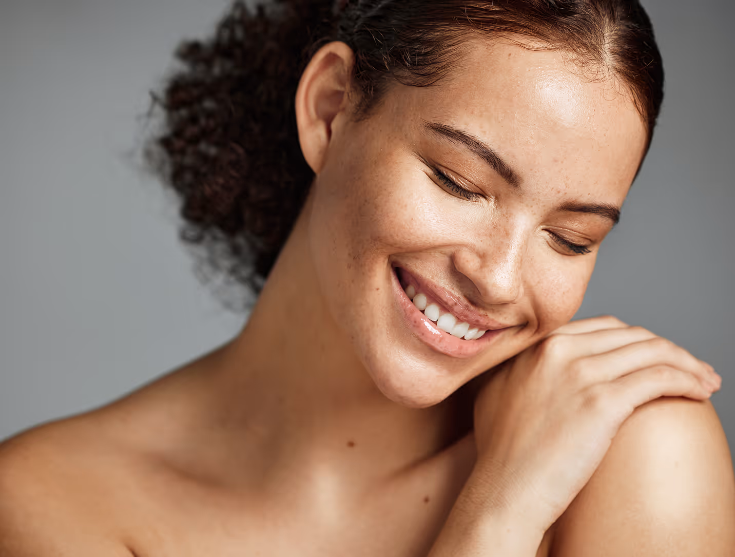 Close-up of a smiling woman with curly hair and clear skin, gently holding her shoulder.