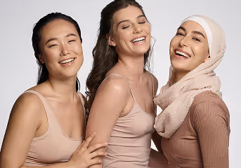 Three diverse women smiling and embracing, wearing light beige and pink tops against a plain background.