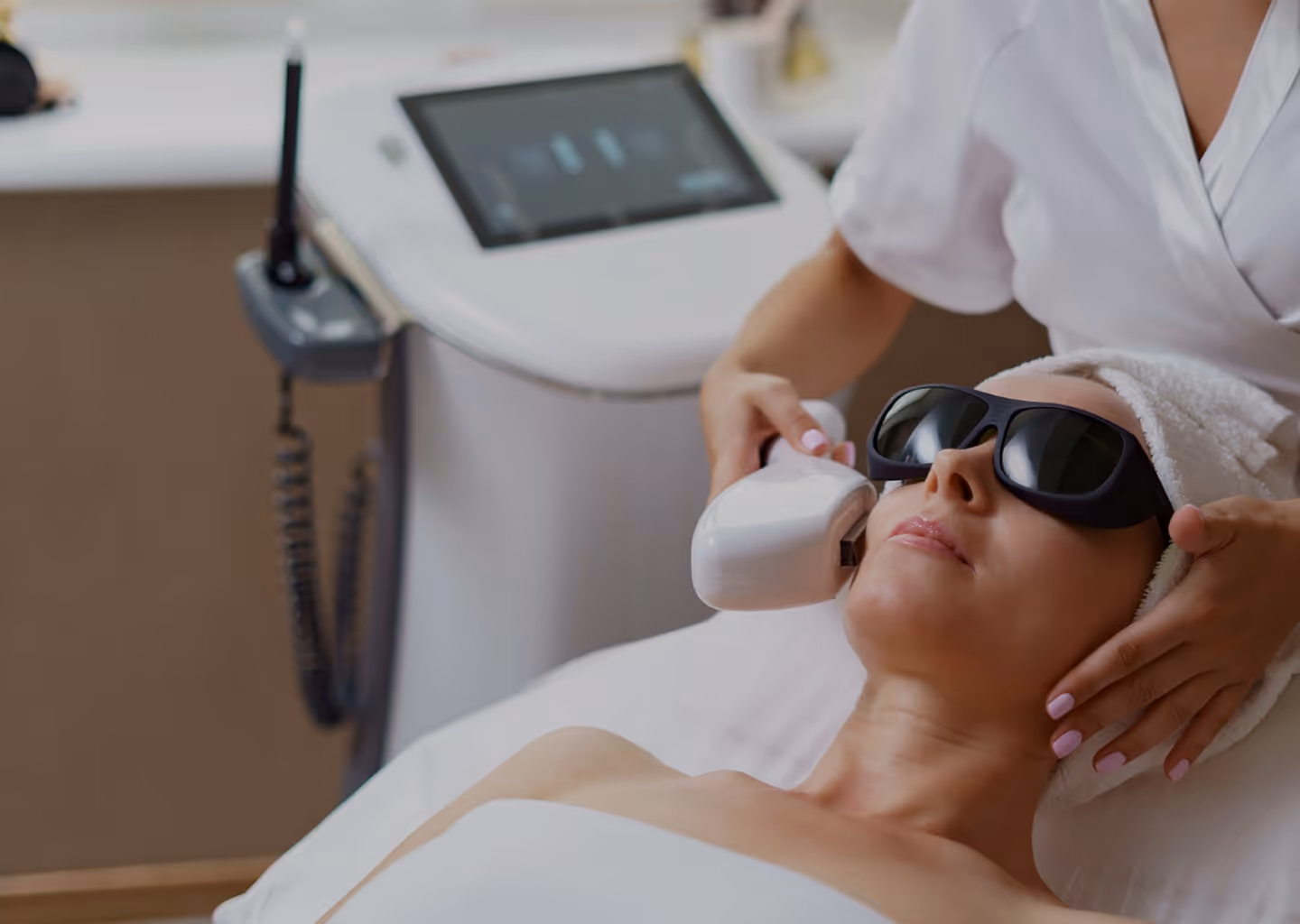 Woman wearing protective glasses receiving a facial laser treatment from a technician in a spa setting.