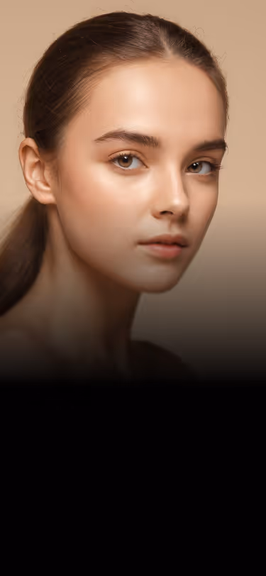 Close-up portrait of a young woman with smooth skin, brown hair pulled back, and light brown eyes against a beige background.