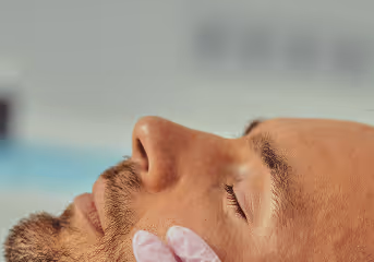 Close-up of a man with eyes closed receiving a skincare treatment with a gloved hand on his cheek.