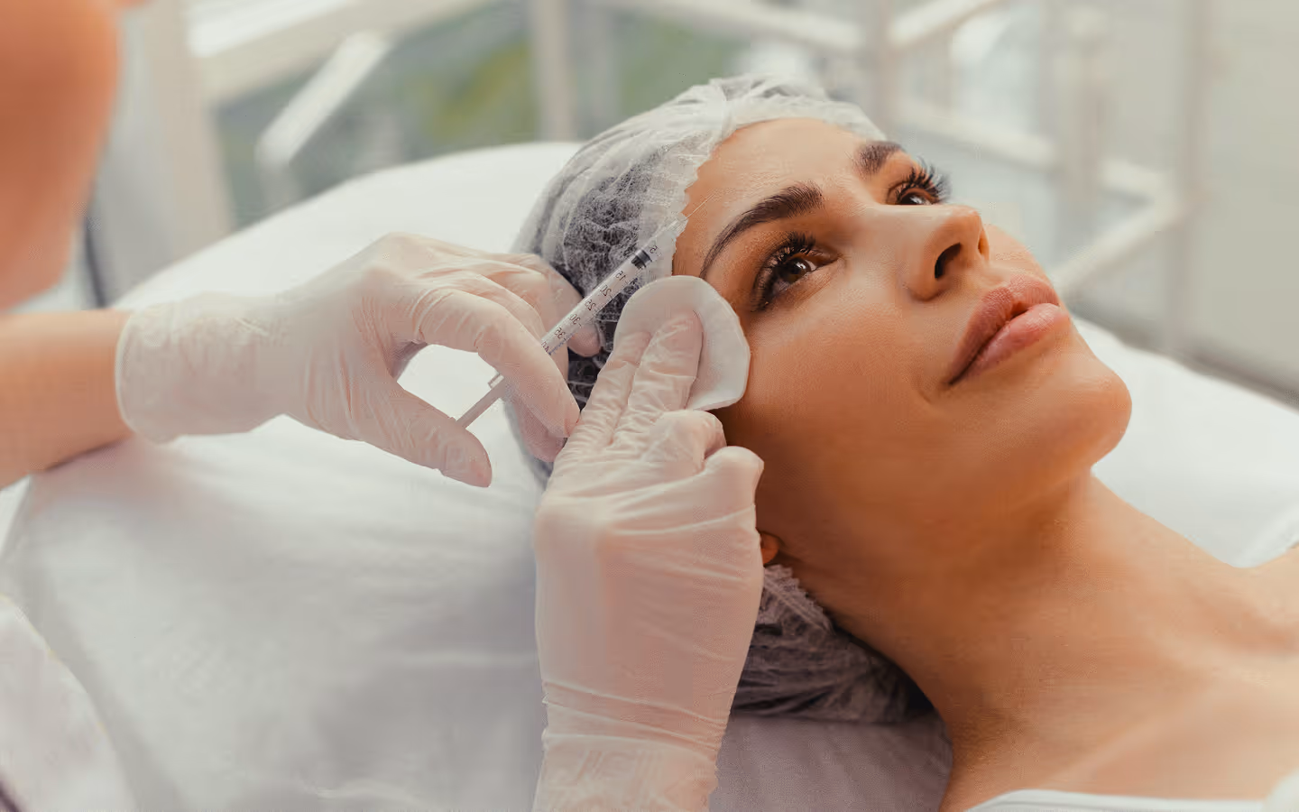 Close-up of a woman receiving a facial injection while wearing a hair cap in a clinical setting.
