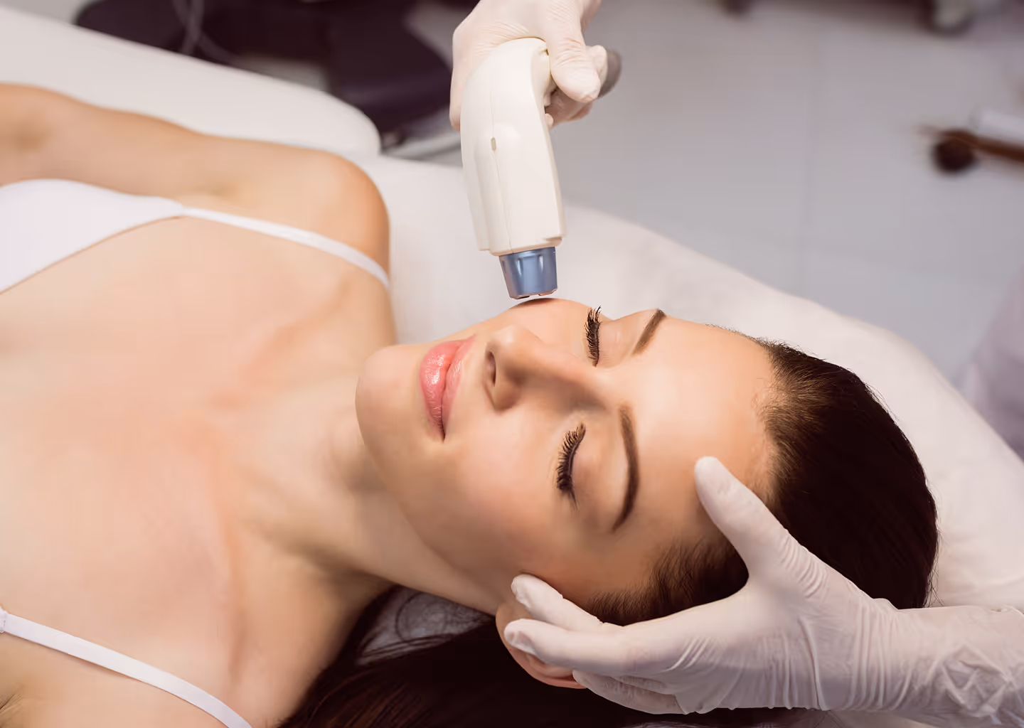 Woman receiving a facial treatment with a handheld device from a gloved dermatologist.