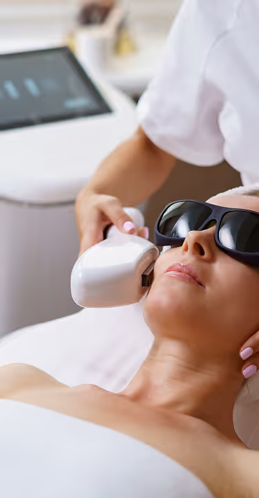 Woman wearing protective glasses receiving facial laser treatment with a handheld device.