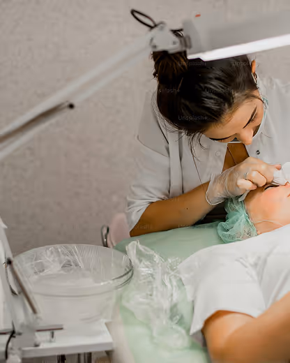 A skincare professional wearing gloves performing a facial treatment on a client lying on a bed.