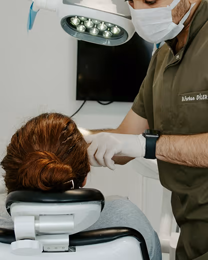 Dentist wearing mask and gloves attending to a patient seated in a dental chair under a dental light.