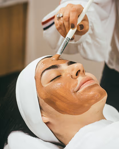A person receiving a brown facial mask applied with a brush while lying with eyes closed and wearing a white headband.