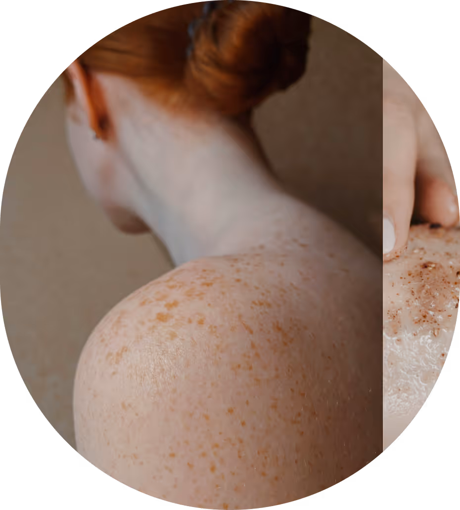 Close-up of a person's freckled shoulder and back with a hand applying a brown exfoliating scrub.