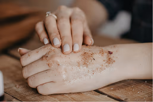 Close-up of a person exfoliating the back of their hand with a brown scrub on a wooden surface.