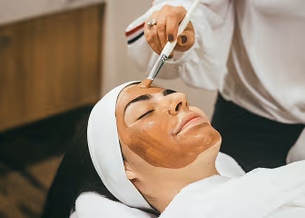 Woman with eyes closed receiving facial treatment with a brush applying a brown mask.