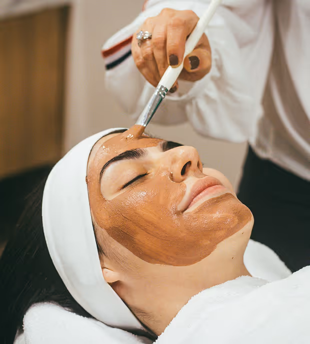 Woman with eyes closed receiving a brown facial mask applied with a brush at a spa.