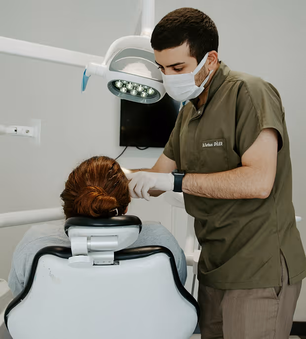 Dentist wearing a mask and gloves examining a patient sitting in a dental chair under an overhead dental light.