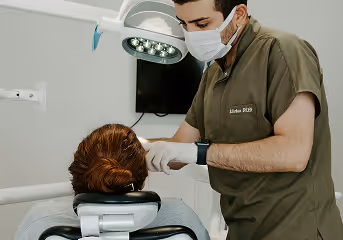 Dentist wearing a surgical mask and gloves examining a patient's teeth under a dental light.