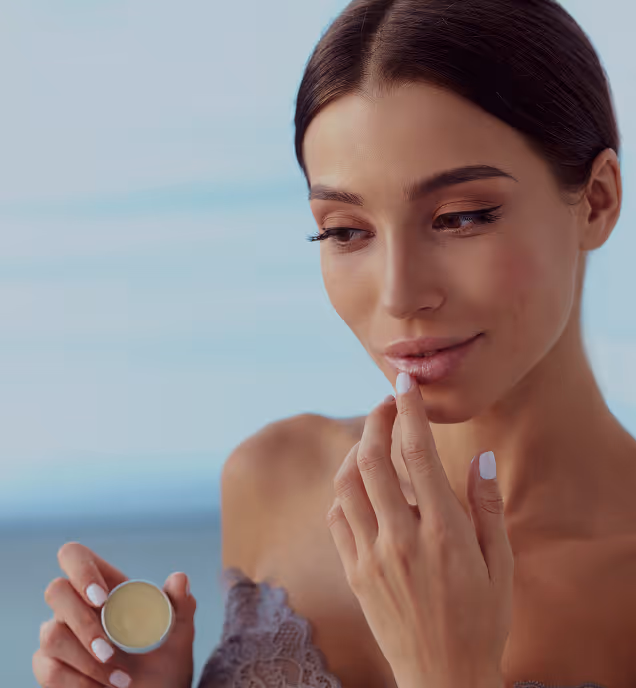 Woman with dark hair applying lip balm from a small container against a soft blue background.