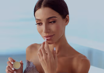 Woman applying lip balm while holding the container in her hand against a blurred background.