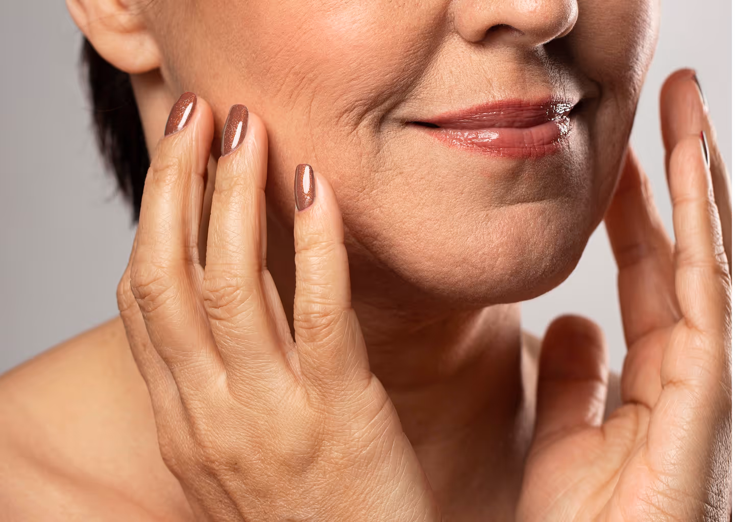 Close-up of a mature woman touching her face with manicured nails and smiling softly.