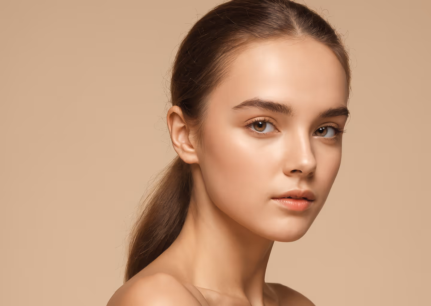 Close-up of a young woman with clear skin and brown hair tied back against a beige background.