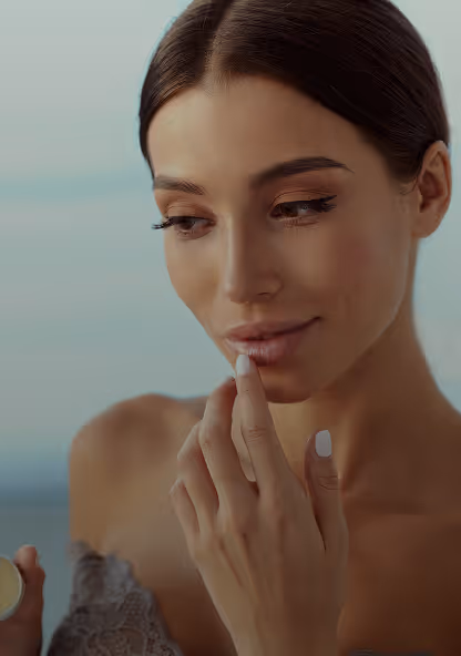 Young woman with smooth skin gently touching her lips while holding a small cosmetic container.