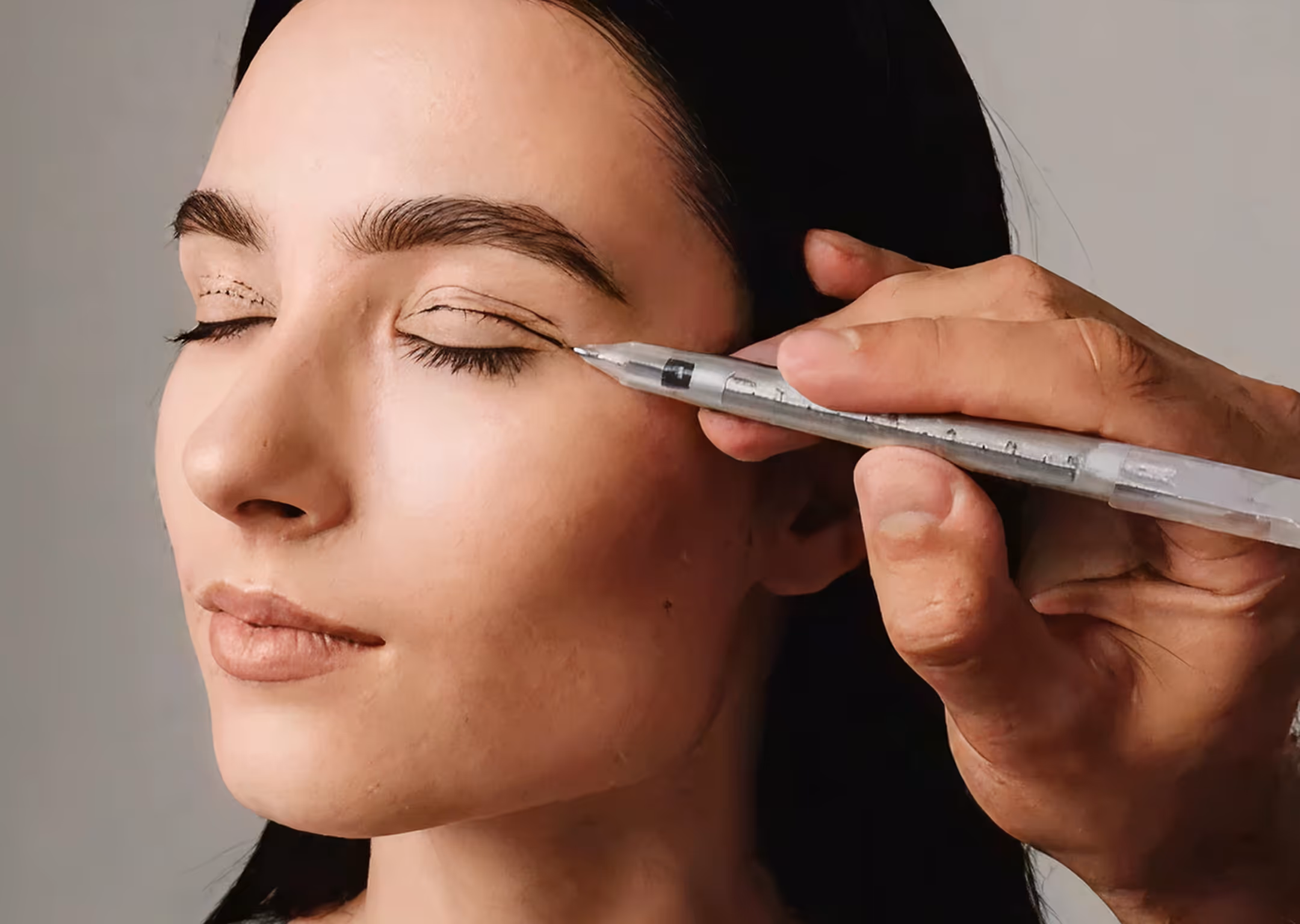 Hand drawing eyeliner lines on closed eyelid of a woman with dark hair.