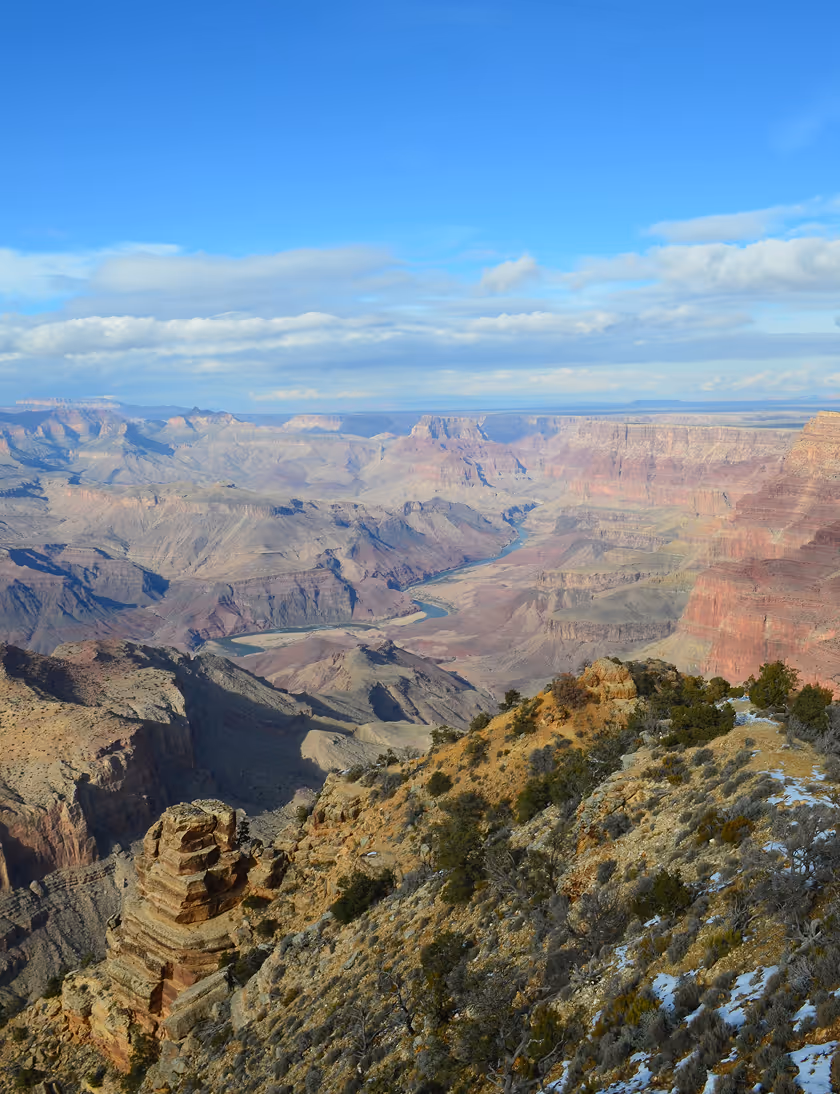 Expansive view of Grand Canyon with winding river and rocky cliffs under a partly cloudy sky.