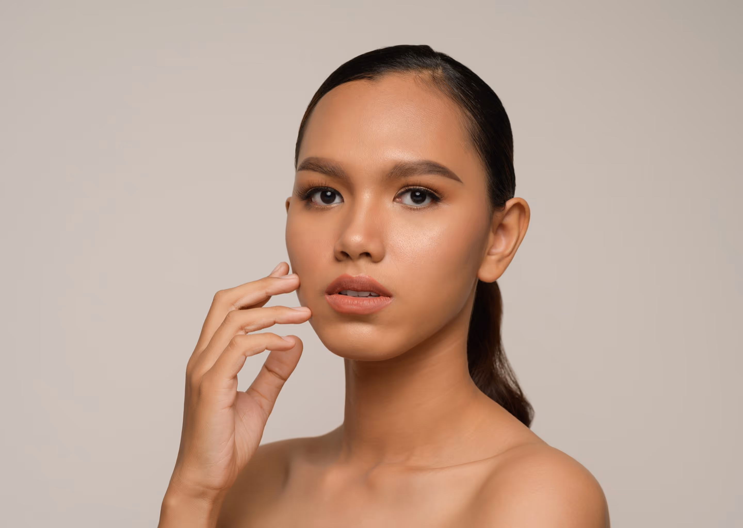 Portrait of a woman with smooth skin and makeup touching her face against a plain background.