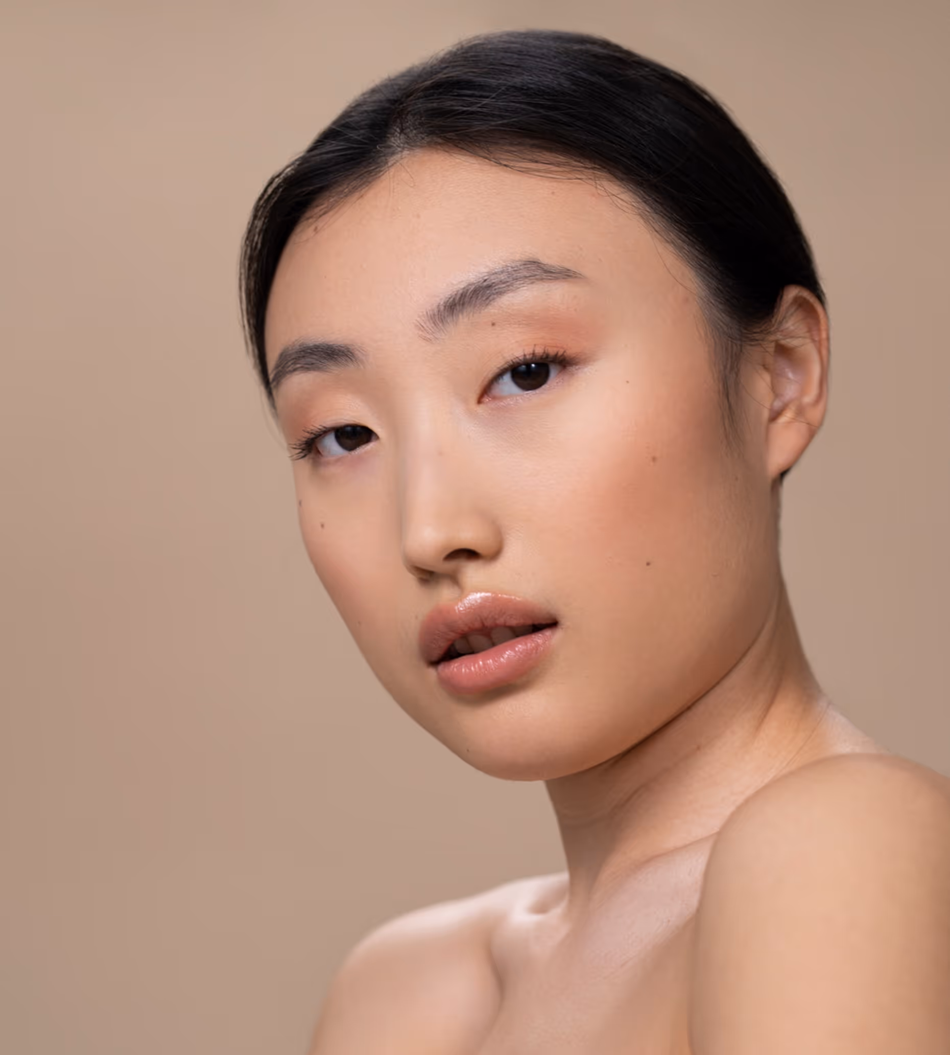 Close-up of a young woman with smooth, natural makeup and glossy lips against a beige background.