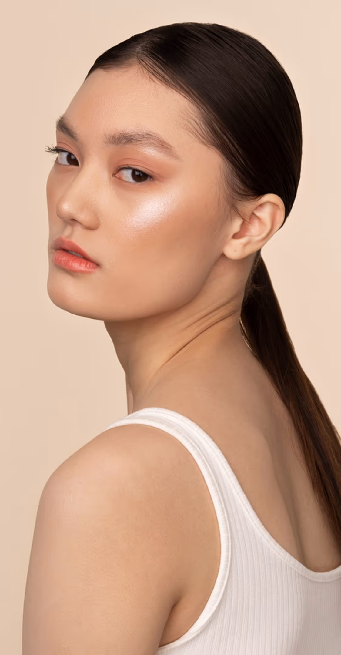 Close-up of a woman with sleek dark hair, glowing skin, and wearing a white tank top against beige.