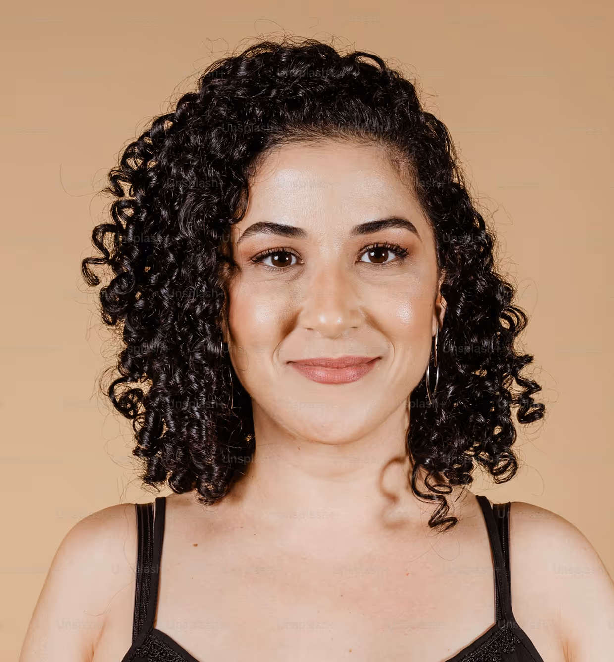Smiling woman with curly black hair wearing hoop earrings and a black top against a beige background.