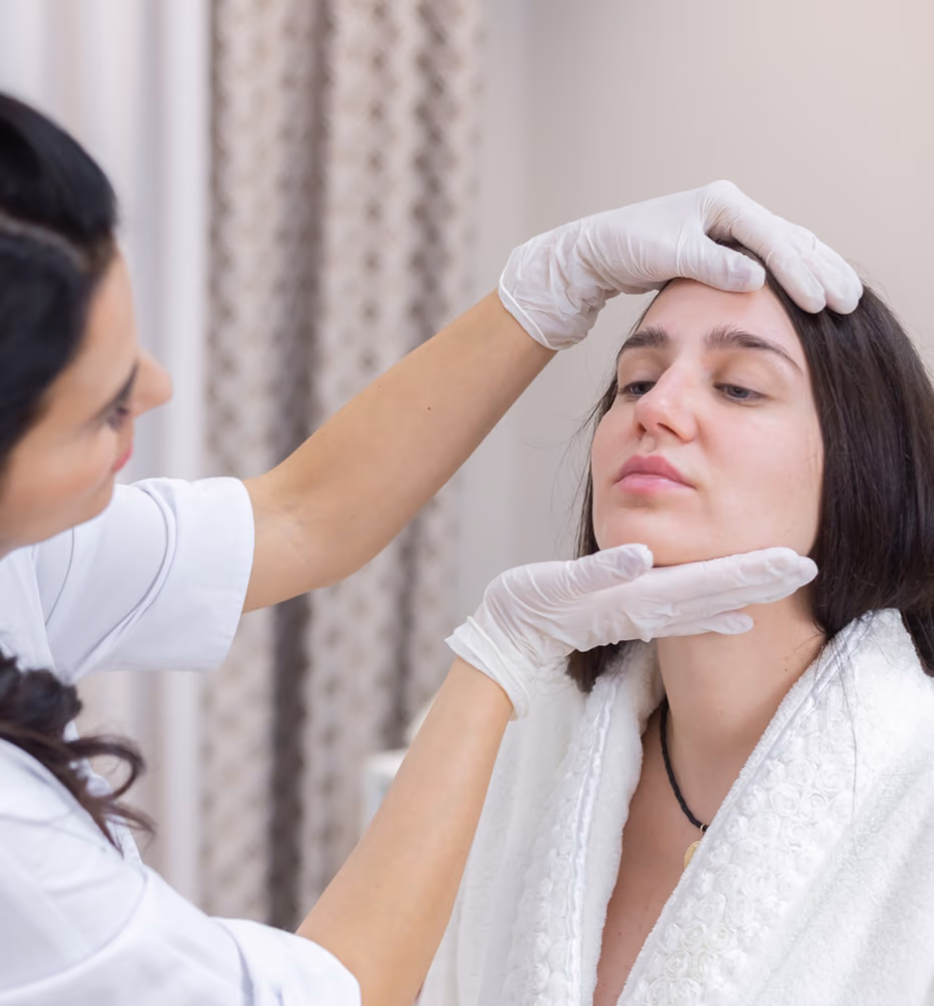 Skincare specialist wearing gloves examines a woman's face in a spa or clinic setting.