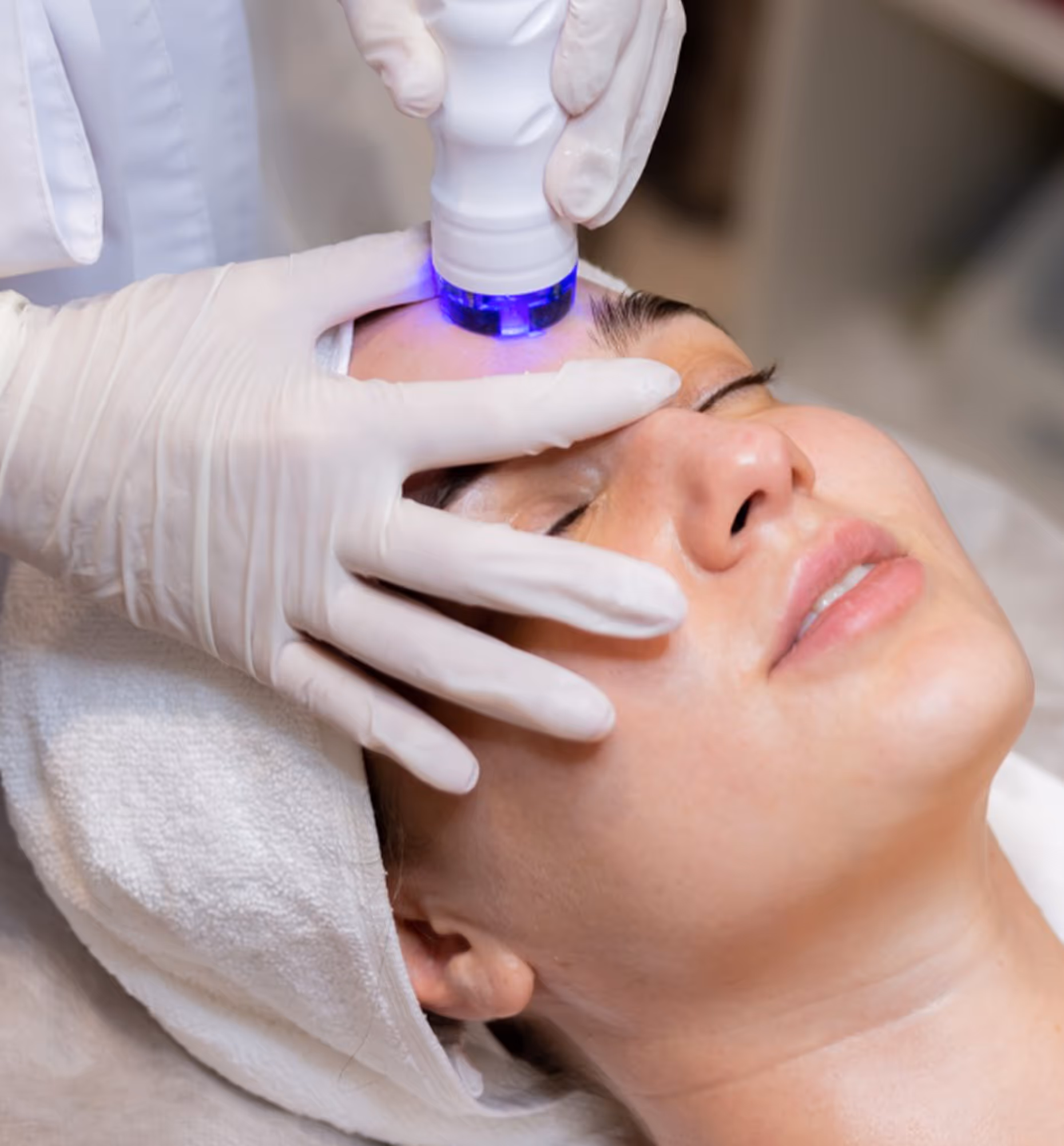 Woman receiving a facial treatment with a blue light device on her forehead by a gloved professional.