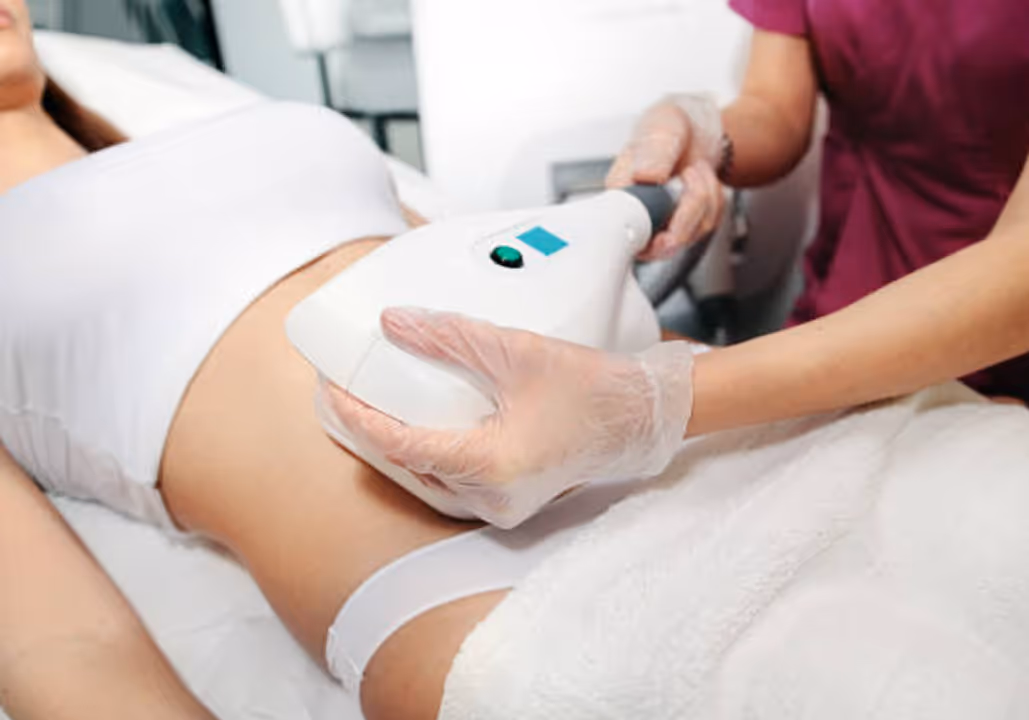 Technician wearing gloves uses a body contouring device on a woman's abdomen lying on a treatment table.