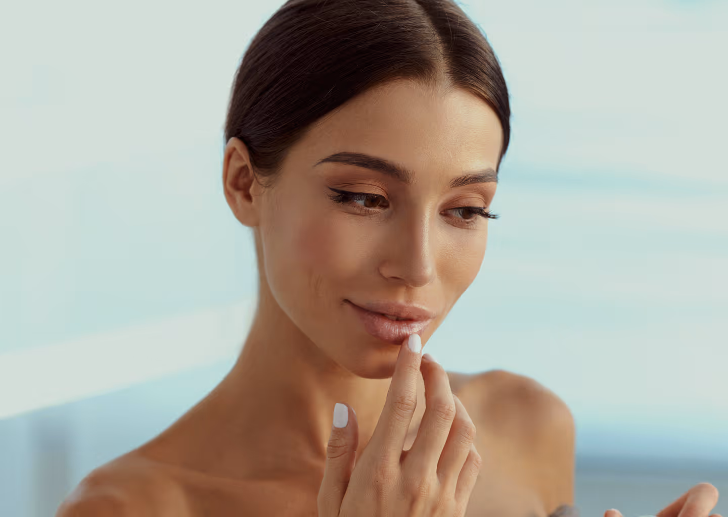 Woman with tied dark hair applying lip balm with finger against a light blue background.