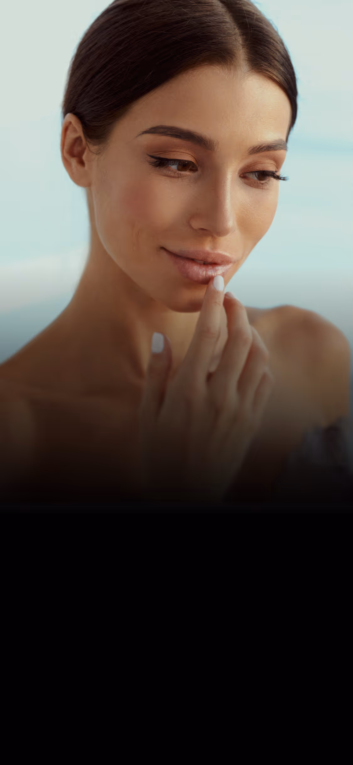 Woman with smooth skin touching her lips while looking downward with a gentle smile.