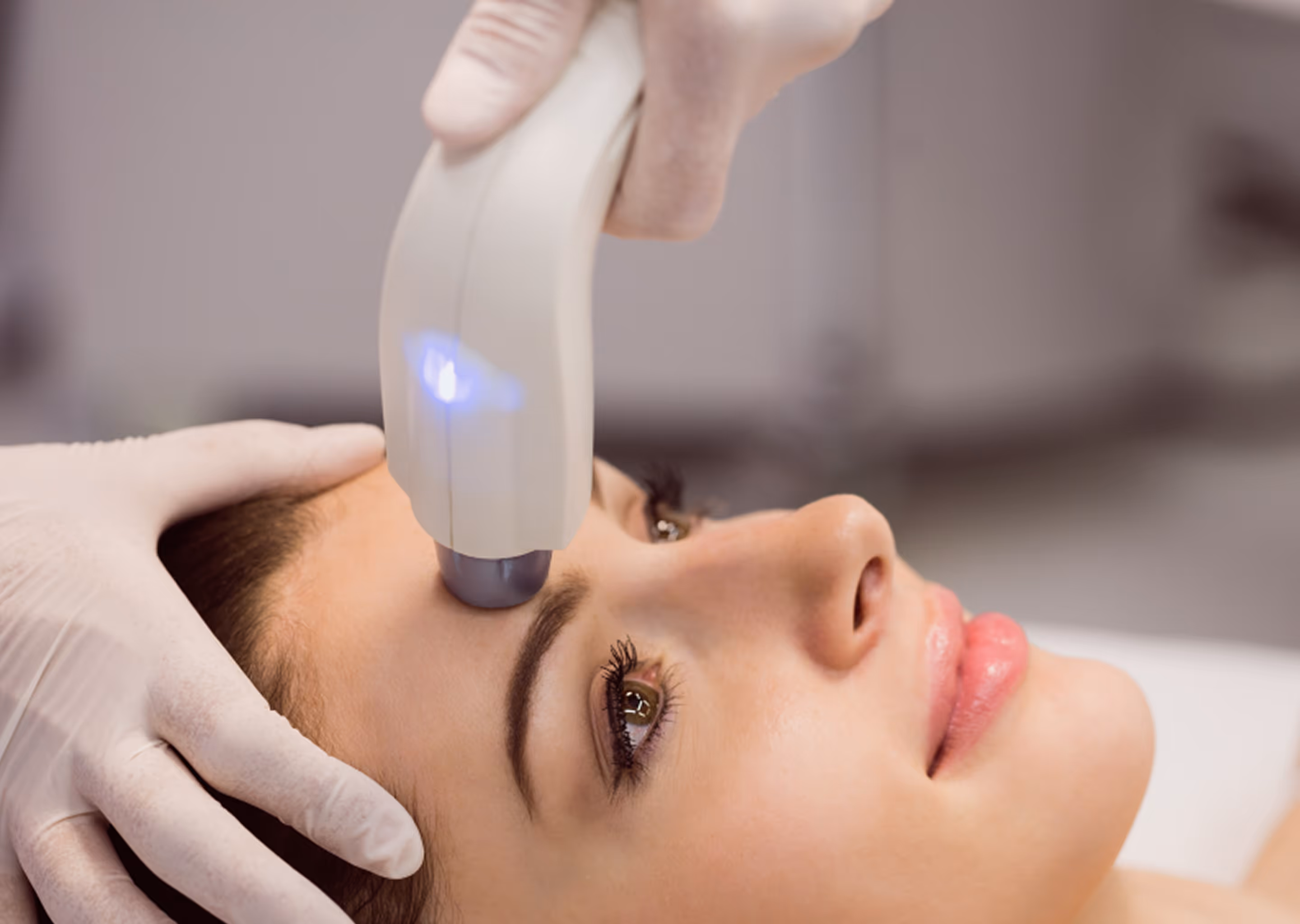 Woman receiving a cosmetic facial treatment with a handheld device emitting blue light on her forehead.